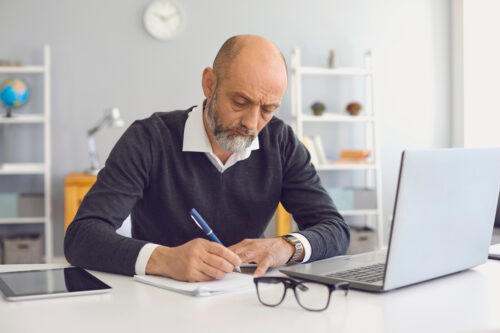 a man taking notes, writing on paper with a pen, glasses and computer on side
