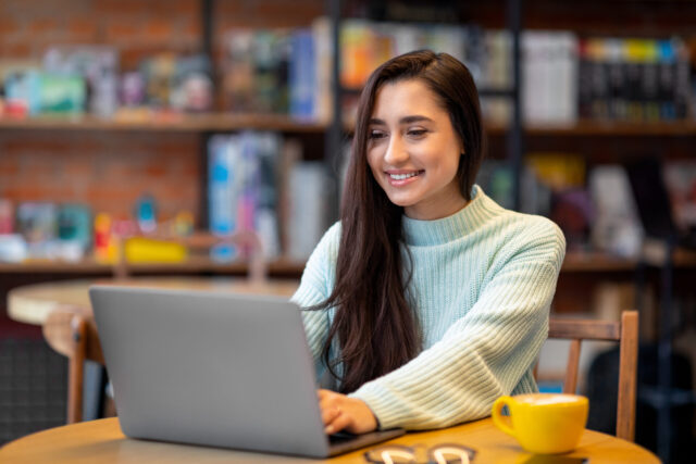 woman working on laptop computer in cafe, looking and smiling at screen, copy space