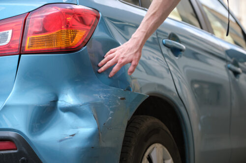 Driver hand examining dented car with damaged fender parked on city street side. Road safety and vehicle insurance concept.